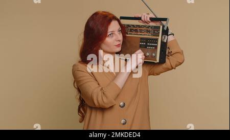 Redhead woman using retro tape record player to listen music, disco dancing of favorite track, having fun, entertaining, fan of vintage technologies. Young ginger girl isolated on beige background Stock Photo