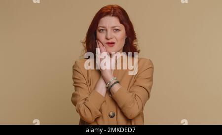 Hipster redhead woman touching sore cheek suffering from toothache cavities or gingivitis waiting for dentist appointment gums disease. Young girl indoor studio shot isolated alone on beige background Stock Photo