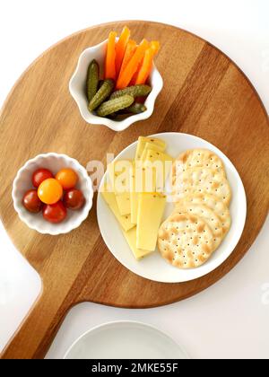 Dutch Gouda cheese slices and water crackers with cherry tomatoes,orange pepper strips and gherkins on round wooden serving board in flat lay composit Stock Photo