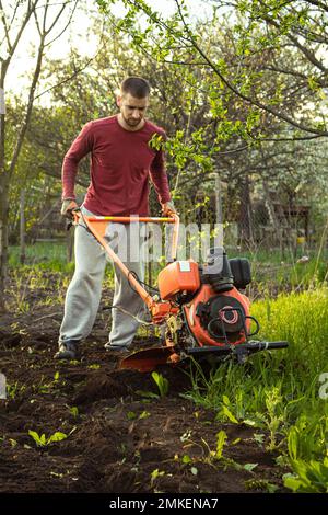 A farmer man works in the field, plowing the land with a plow on the farm. A plowman on a walk behind a motor cultivator. Tillage season. Organic cult Stock Photo
