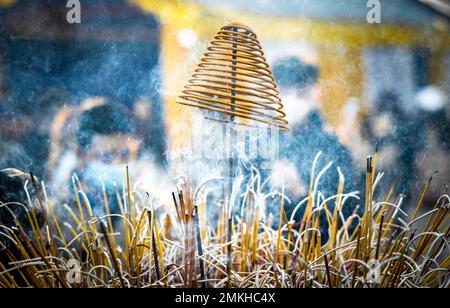 A spiral incense burns above normal incense sticks generating clouds of smoke as worshippers pray behind at Tran Quoc Pagoda in Hanoi, Vietnam. Stock Photo