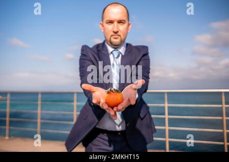 Man persimmon park. Hipster millennial man in a tie and jacket bites off a persimmon while sitting on a park bench in slow motion Young business Stock Photo