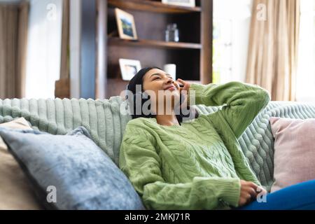 Image of happy biracial woman in headphones, relaxing on sofa with eyes closed at home, copy space. Leisure, relaxation, technology and domestic life Stock Photo
