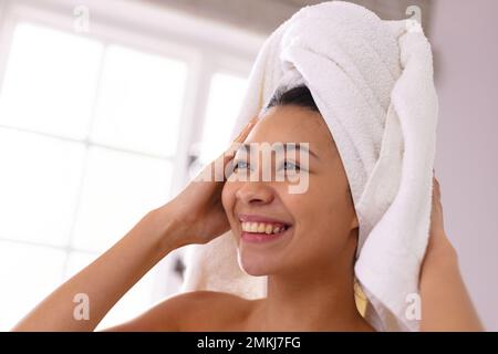 Happy biracial woman wearing towel on head smiling in bathroom, with copy space. Relaxation, self care, beauty, leisure and lifestyle concept. Stock Photo