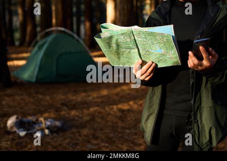 Adult hiker with backpack using mobile phone and map in forest near his tent. Hiking elderly man in autumn nature holding map outdoors next to a Stock Photo