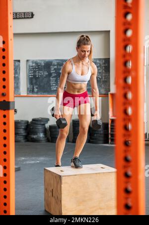 Full body of strong female doing step up exercise with dumbbells on wooden box during intense functional workout in gym Stock Photo