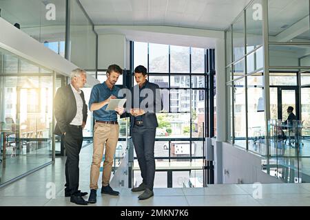 Three eyes on the project. three colleagues talking together over a digital tablet while standing in a modern office. Stock Photo