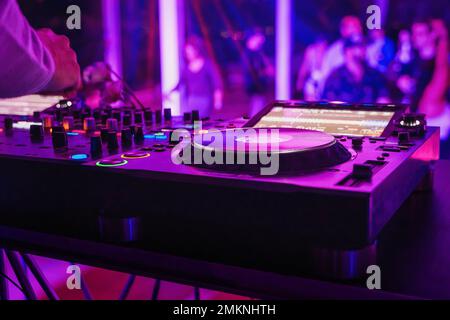Close-up of a DJ booth at a nightclub, with the focus on one of the turntables and the colorful lighting of the dance floor Stock Photo