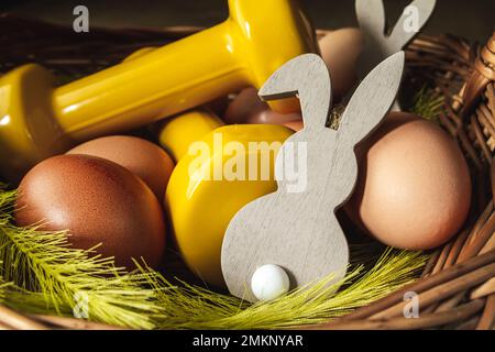 Traditional Easter wicker basket with eggs, decorative bunny and dumbbells. Healthy fitness diet choice. Gym workout, dieting training concept. Stock Photo