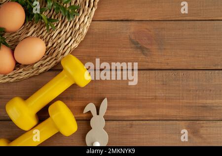 Gym dumbbells, decorative Easter bunny, eggs and boxwood branches. Healthy fitness workout flat lay, training concept with copy space on wood. Stock Photo