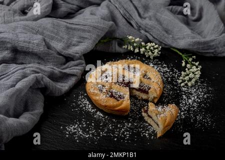 A small chocolate tart covered in icing sugar with white flowers and a gray scarf next to it, on a black slate background Stock Photo