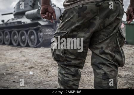 Soldier legs on a battlefield, wearing woodland camo military pants, camouflage trousers. Army battle tank in background. Stock Photo