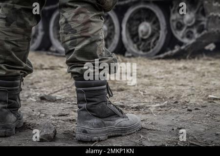 Soldier legs on a battlefield, wearing woodland camo military pants, camouflage trousers. Army tank tracked wheels in background. Stock Photo