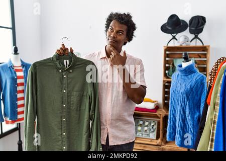 African man with curly hair holding shirt from clothing rack at retail shop serious face thinking about question with hand on chin, thoughtful about c Stock Photo