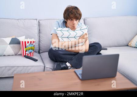 Hispanic young man watching a horror movie in the laptop making fish face with mouth and squinting eyes, crazy and comical. Stock Photo
