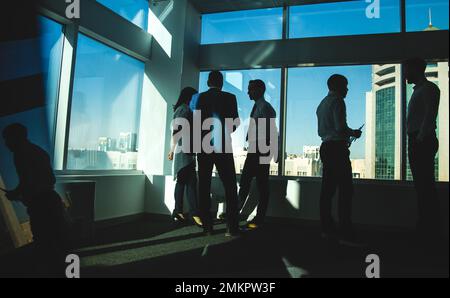 Silhouettes of people standing near a panoramic window in a modern office. Team of young professional business people working and chatting together in Stock Photo