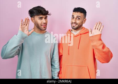 Young hispanic gay couple standing over pink background waiving saying hello happy and smiling, friendly welcome gesture Stock Photo