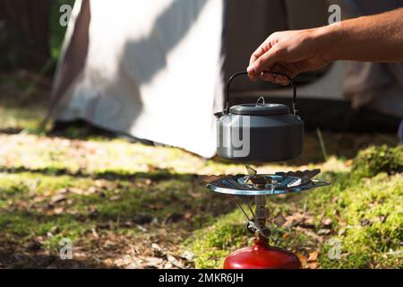 Cooking, heating a tourist kettle on a portable gas burner with a red gas cylinder. Camping, a man cooks breakfast outdoors. Summer outdoor activities Stock Photo