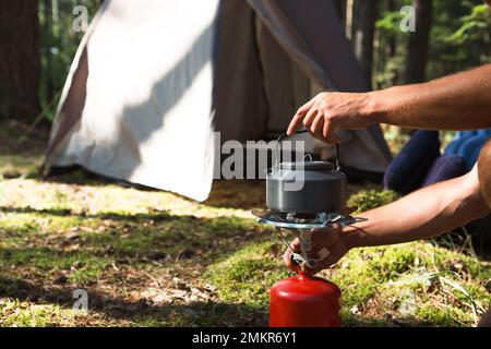 Cooking, heating a tourist kettle on a portable gas burner with a red gas cylinder. Camping, a man cooks breakfast outdoors. Summer outdoor activities Stock Photo