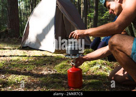 Cooking, heating a tourist kettle on a portable gas burner with a red gas cylinder. Camping, a man cooks breakfast outdoors. Summer outdoor activities Stock Photo