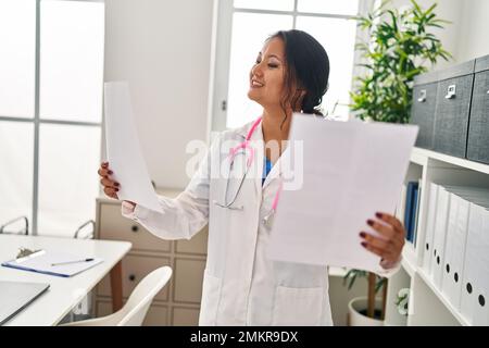 Young chinese woman wearing doctor uniform reading document at clinic Stock Photo