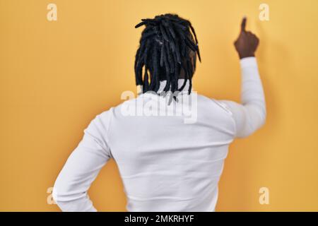 African man with dreadlocks wearing turtleneck sweater over yellow background posing backwards pointing ahead with finger hand Stock Photo