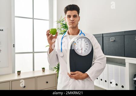 Young hispanic doctor man holding scale at dietitian clinic puffing cheeks with funny face. mouth inflated with air, catching air. Stock Photo