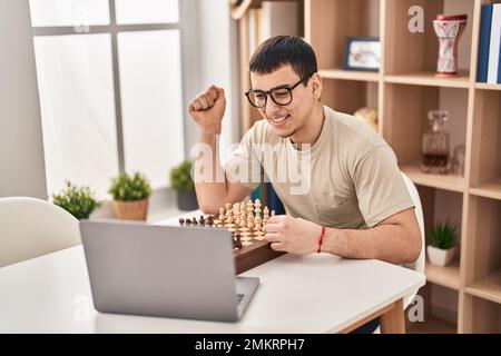 Young arab man playing chess online screaming proud, celebrating victory and success very excited with raised arm Stock Photo