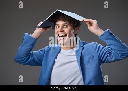 happy caucasian young guy in blue jacket holding laptop computer over head isolated on gray background. Stock Photo