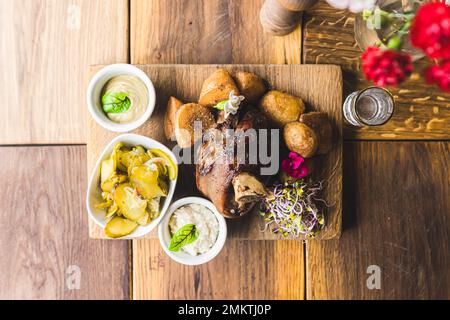 Top-down view of roasted ham hock served with golden potatoes horseradish mustard and pickles on wooden board. Polish cuisine. Horizontal indoor shot. High quality photo Stock Photo