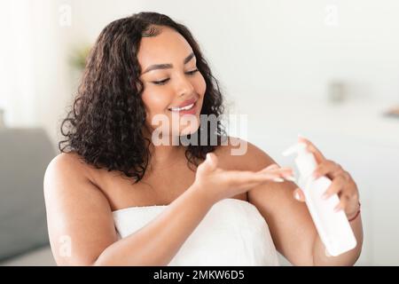 Black oversize woman holding bottle and pouring moisturising body lotion on hand, using new skincare product after bath Stock Photo