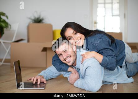 Happy Young Spouses Embracing And Using Laptop After Moving To Their New Home, Stock Photo