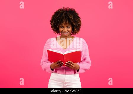 Cheerful smart pretty young black curly female student in casual reading book, diary, plans day with notepad Stock Photo