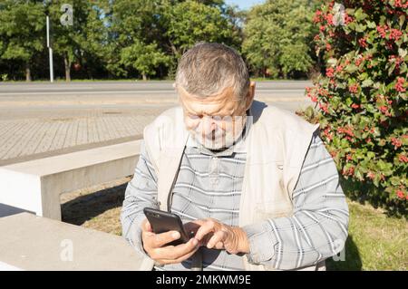Caucasian senior man sitting on  bench and using cellular at sunny autumnal day in Ukraine Stock Photo