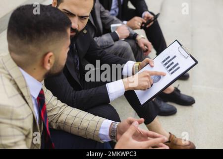 Business people outdoor meeting. A company of male businessmen in suits are sitting on the steps of the stairs. Working break. Teamwork and Stock Photo