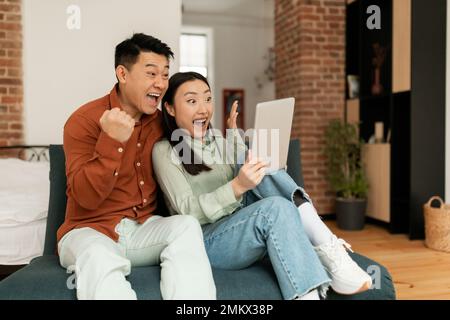 Surprised asian spouses using digital tablet, looking at screen with excitement and clenching fists, sitting on sofa Stock Photo