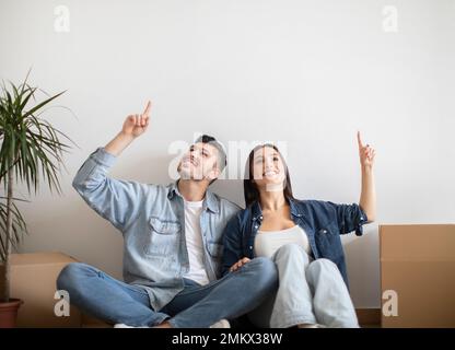 Happy Young Spouses Sitting On Floor After Moving And Pointing Up Stock Photo