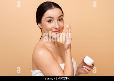 Beautiful Young Indian Female Applying Moisturising Cream On Face Over Beige Background Stock Photo