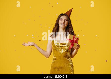 Happy young lady in birthday hat and dress holding gift box, standing over yellow background with falling confetti Stock Photo