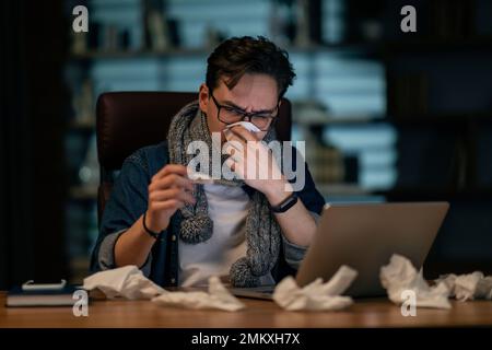 Sick young man sitting in front of computer, holding thermometer Stock Photo