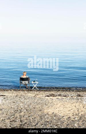 A blonde woman sits on a camping chair on the shore of Kattegatt in the sun and drinks wine, Djursland, Jutland, Denmark Stock Photo