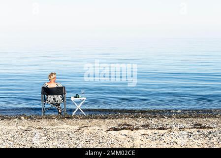 A blonde woman sits on a camping chair on the shore of Kattegatt in the sun and drinks wine, Djursland, Jutland, Denmark Stock Photo