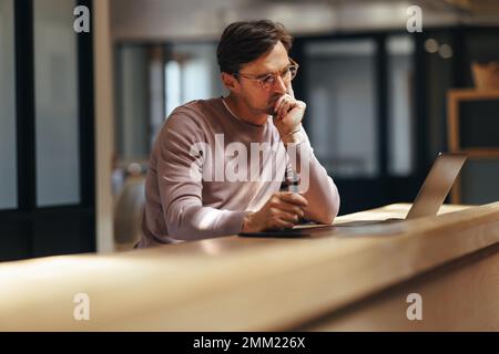 Pensive web designer working on a project in a coworking office. Male designer thinking and looking at his design on a laptop. Creative business man w Stock Photo
