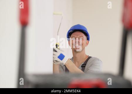Happy painter in hard hat paints wall with white paint and looks at camera. Stock Photo