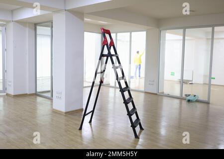 Metal ladder standing in center of empty office, worker repairing wall in background. Stock Photo