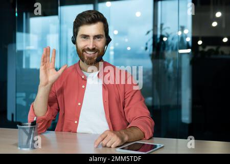 Portrait of a young man in a red shirt and headset. He sits at the desk in the office, looks at the camera, waves his hand, smiles. Stock Photo