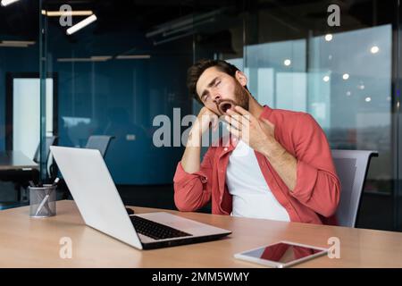 A tired young man in a red shirt sits at the desk in the office and yawns, covering his mouth with his hand. Works overtime, boring monotonous work, falls asleep. Stock Photo