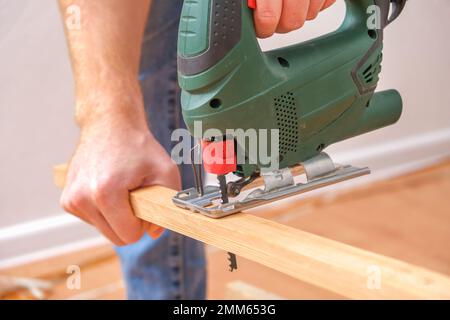 The hands of a master carpenter with an electric jigsaw in his hands cutting off a piece of wood. Male hands using fret saw for cutting wood. A carpenter cuts wood with a jigsaw in a home workshop. Stock Photo