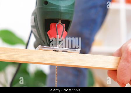 The hands of a master carpenter with an electric jigsaw in his hands cutting off a piece of wood. Male hands using fret saw for cutting wood. A carpenter cuts wood with a jigsaw in a home workshop. Stock Photo