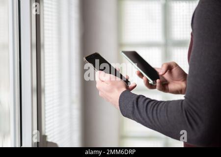Close up of a man holding two smartphones. Man using two phones at the same time.Busy man standing next to windows and using two smartphones. Stock Photo